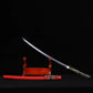 Japanese katana Sakura no Ken displayed diagonally on a wooden stand, black background, unsheathed black steel blade with engraved sakura floral pattern, curved blade profile visible, red leather wrapped saya placed horizontally below, black and white tsuka handle, traditional samurai sword presentation
