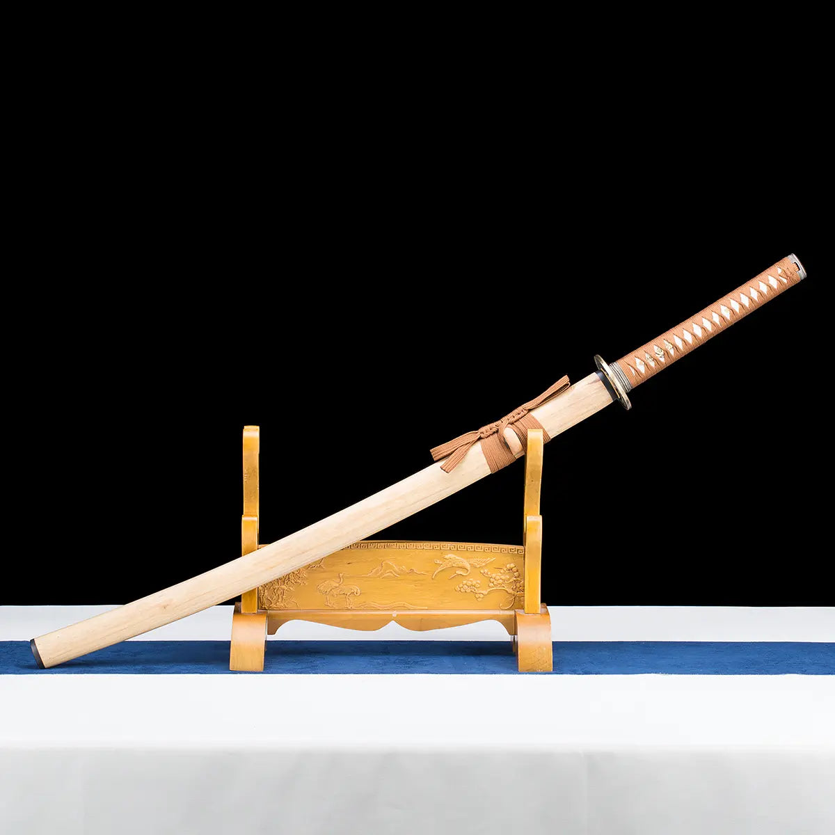 Angled display view of the Japanese artisanal katana Take no Kaze photographed on a deep black background, presented on a wooden stand. The katana is shown sheathed, with the natural light bamboo scabbard resting diagonally across the stand. The brown woven sageo is tied neatly around the saya, while the handle wrapped in brown synthetic leather ito over white ray skin extends upward. The round dark metal tsuba and brass-colored habaki are visible near the scabbard opening, emphasizing the traditional mount