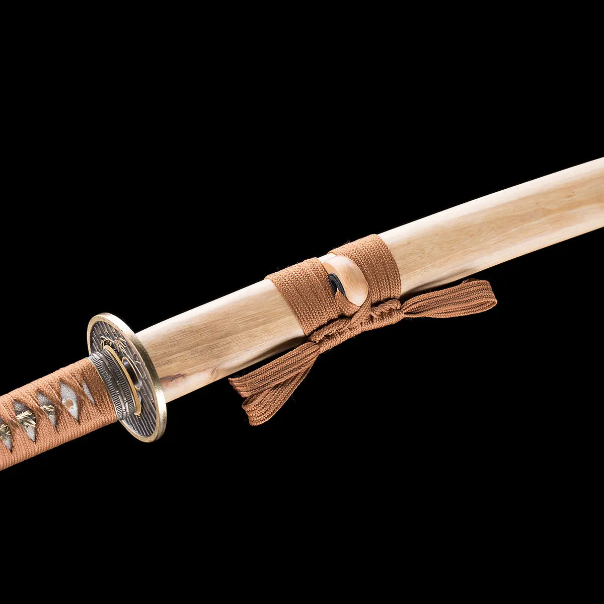 Detail view of the Japanese artisanal katana Take no Kaze photographed on a solid black background, showing the bamboo scabbard with a natural light wood grain finish. The brown woven sageo is neatly tied around the saya, with the kurigata clearly visible. Part of the handle wrapped in brown synthetic leather ito and the circular dark metal tsuba can be seen near the scabbard opening, highlighting the traditional katana mounting and natural bamboo construction.

