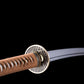 Detailed close-up of the Japanese artisanal katana Take no Kaze photographed on a deep black background, focusing on the round metal tsuba decorated with engraved bamboo motifs. The guard shows a dark textured surface with bamboo leaf details and a brass-colored rim. Part of the handle wrapped in brown synthetic leather ito over white ray skin is visible, along with the polished steel blade extending outward, highlighting the traditional katana mounting and bamboo-inspired design.
