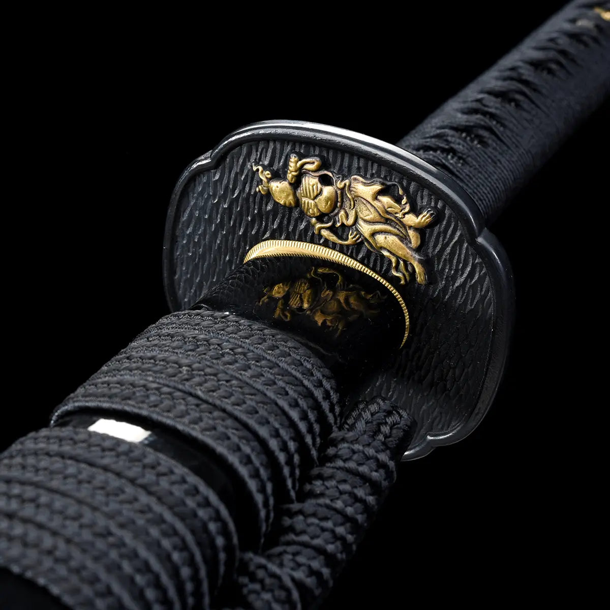 Close-up view of a Japanese katana guard photographed on a deep black background, highlighting the black textured tsuba adorned with a gold dragon motif, fine engraved details clearly visible, black tsuka wrapped in traditional black ito, gold habaki at the base of the blade, emphasizing high-end craftsmanship, symbolic design, and premium samurai sword fittings.
