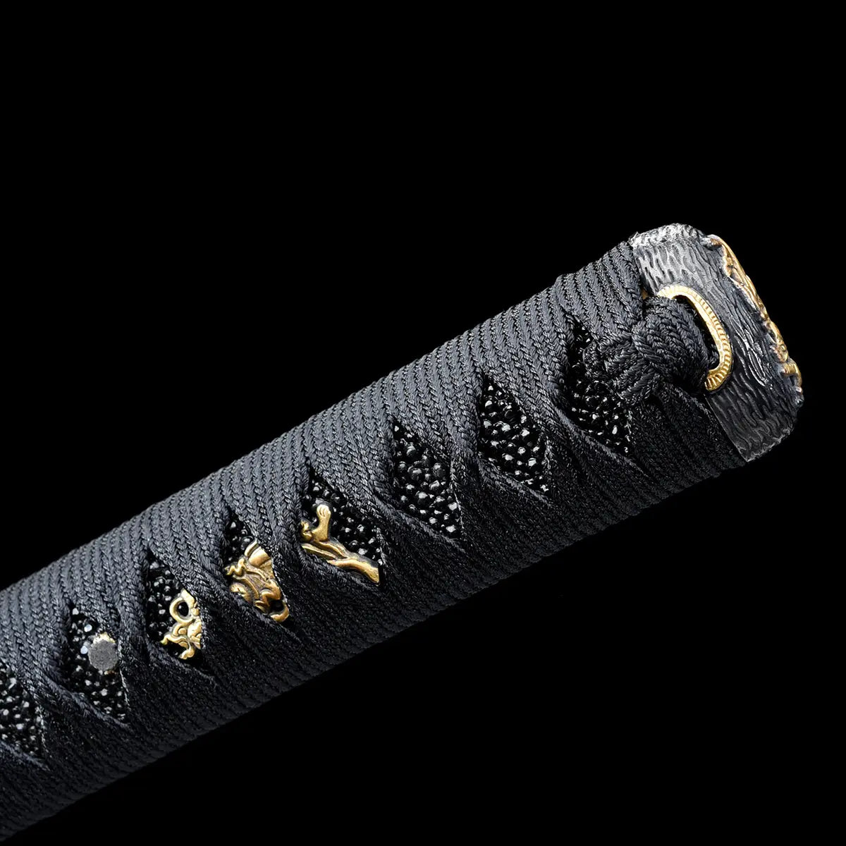 Detailed close-up of a Japanese katana handle photographed on a deep black background, showing the traditional black tsuka wrapped with black ito, textured black ray skin underneath, and gold menuki ornaments embedded beneath the wrap, with a gold kashira ring visible at the pommel, emphasizing premium craftsmanship, traditional assembly, and refined samurai sword aesthetics.
