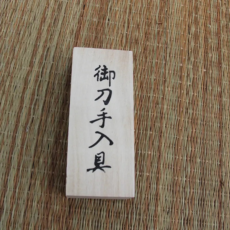 Couvercle de coffret en bois clair avec kanji noirs, posé sur tatami tressé beige/brun, vue de dessus, lumière douce — coffret de rangement du kit d’entretien katana, accessoires de maintenance pour sabre japonais.
