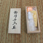 Coffret en bois ouvert posé sur tatami beige/brun, contenu visible sous sachets : flacon d’huile chōji, papier de riz jaune, boule d’uchiko orange ; couvercle avec kanji à gauche, vue de dessus, lumière naturelle — kit d’entretien katana, accessoires de maintenance pour sabre japonais.