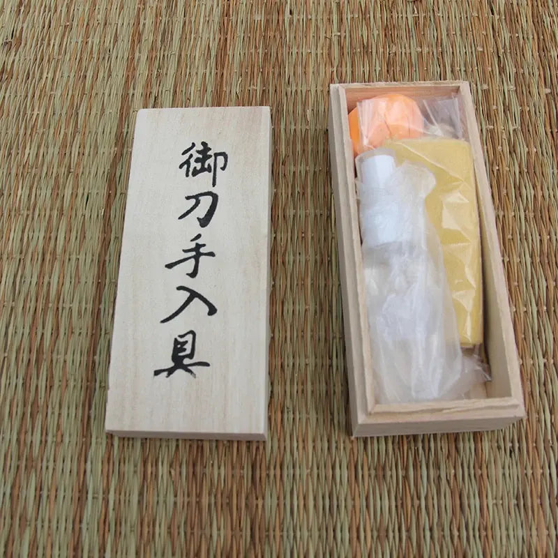 Coffret en bois ouvert posé sur tatami beige/brun, contenu visible sous sachets : flacon d’huile chōji, papier de riz jaune, boule d’uchiko orange ; couvercle avec kanji à gauche, vue de dessus, lumière naturelle — kit d’entretien katana, accessoires de maintenance pour sabre japonais.