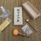 Vue d’ensemble du kit d’entretien pour katana posé sur tatami beige/brun : flacon spray d’huile chōji, papier de riz jaune, boule d’uchiko orange sur manche bois, mekugi-nuki en laiton, sachet de mekugi en bambou, coffret en bois ouvert avec couvercle à kanji — kit entretien katana, accessoires maintenance sabre japonais.