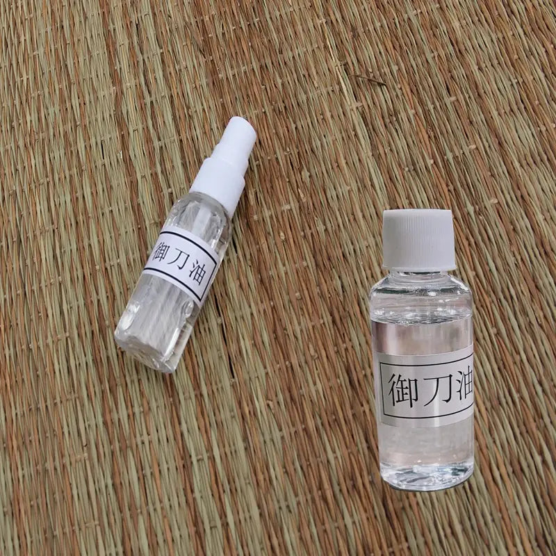 Deux flacons d’huile chōji pour katana sur tatami tressé beige/brun : un spray incliné à gauche et un flacon à bouchon blanc à droite, reflets clairs — kit entretien katana, huile de protection lame, accessoires maintenance sabre japonais.