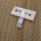 Flacon spray d’huile chōji transparent posé en biais devant le couvercle en bois clair marqué de kanji, sur tatami tressé beige/brun, lumière naturelle — kit d’entretien katana, huile de protection lame, accessoires de maintenance pour sabre japonais.