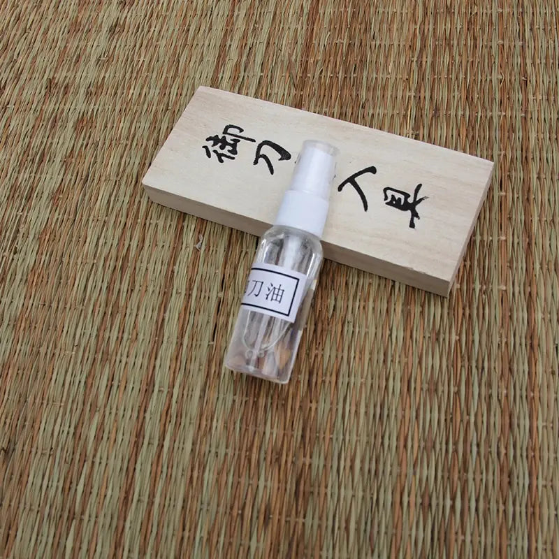 Flacon spray d’huile chōji transparent posé en biais devant le couvercle en bois clair marqué de kanji, sur tatami tressé beige/brun, lumière naturelle — kit d’entretien katana, huile de protection lame, accessoires de maintenance pour sabre japonais.