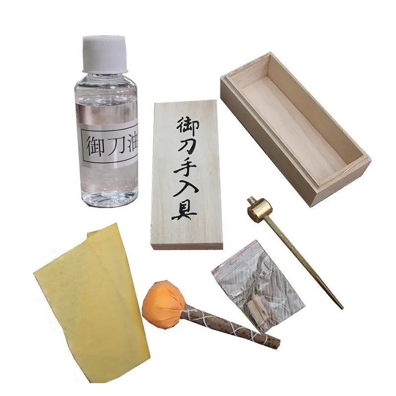 Vue à plat du kit d’entretien pour katana sur fond blanc : flacon d’huile choji transparent, boule d’uchiko orange sur manche bois, papier de riz jaune, mekugi-nuki en laiton, sachet de mekugi en bambou, coffret en bois ouvert avec couvercle à kanji — kit entretien katana, accessoires de maintenance pour sabre japonais.