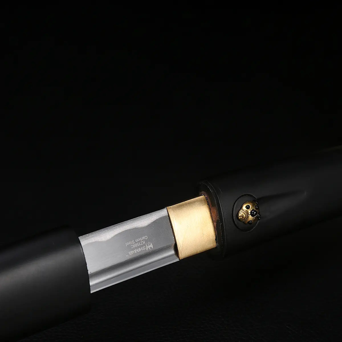 Détail de la jonction lame et poignée du katana shirasaya noir KUROTAKE sur fond noir, montrant l’habaki en laiton, la lame en acier poli partiellement sortie du fourreau, marquage discret gravé sur la lame, poignée et saya en bois dur laqué noir imitation bambou, finition mate sans tsuba.
