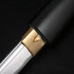 Gros plan sur l’habaki en laiton du katana shirasaya noir KUROTAKE, photographié sur fond noir texturé, montrant la jonction précise entre la lame en acier poli et le fourreau en bois dur laqué noir imitation bambou, finition mate, détails de surface visibles, ensemble sans tsuba.
