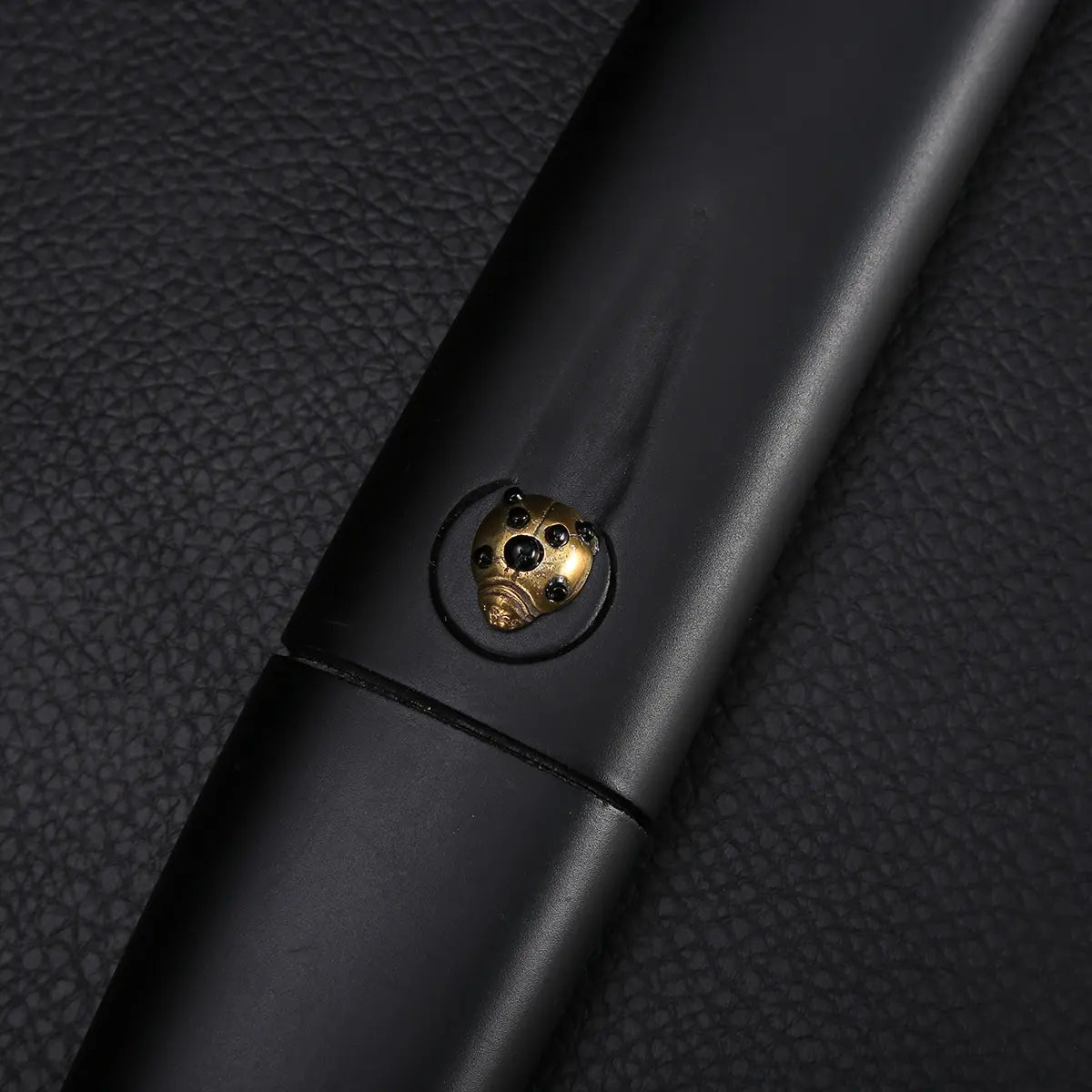 Gros plan sur la poignée du katana shirasaya noir KUROTAKE, photographié sur fond noir texturé, montrant le bois dur laqué noir imitation bambou et un insert décoratif en laiton en forme de tête stylisée, finition mate, jonction nette entre poignée et fourreau, ensemble sans tsuba.
