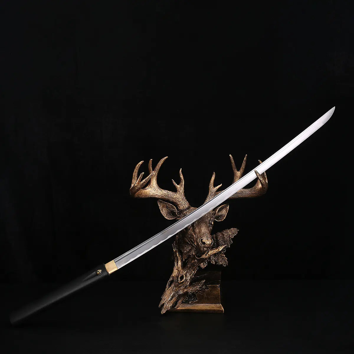 Katana shirasaya noir KUROTAKE avec la lame sortie du fourreau, présenté en diagonale sur un support décoratif en forme de tête de cerf, fond noir profond, lame en acier poli visible sur toute sa longueur avec ligne de trempe discrète, poignée et fourreau en bois dur laqué noir imitation bambou, habaki en laiton apparent, ensemble sans tsuba.
