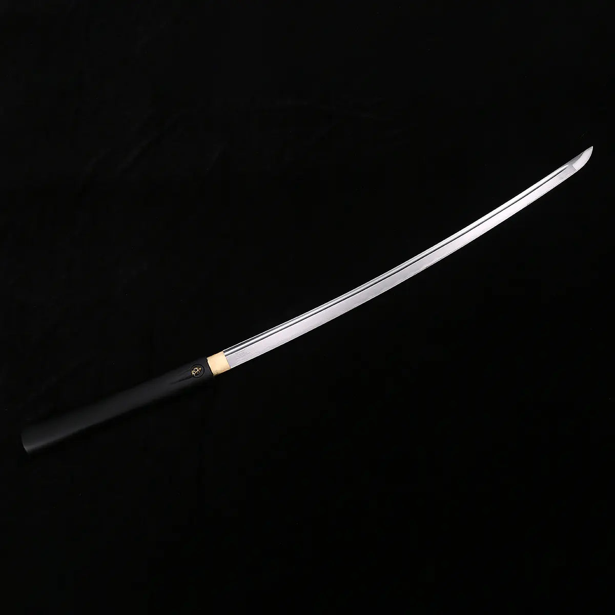 Vue complète de la lame du katana shirasaya noir KUROTAKE photographiée en diagonale sur fond noir, montrant l’acier poli sur toute la longueur avec courbure harmonieuse, pointe marquée visible, habaki en laiton et poignée en bois dur laqué noir imitation bambou partiellement visibles, ensemble sans tsuba.
