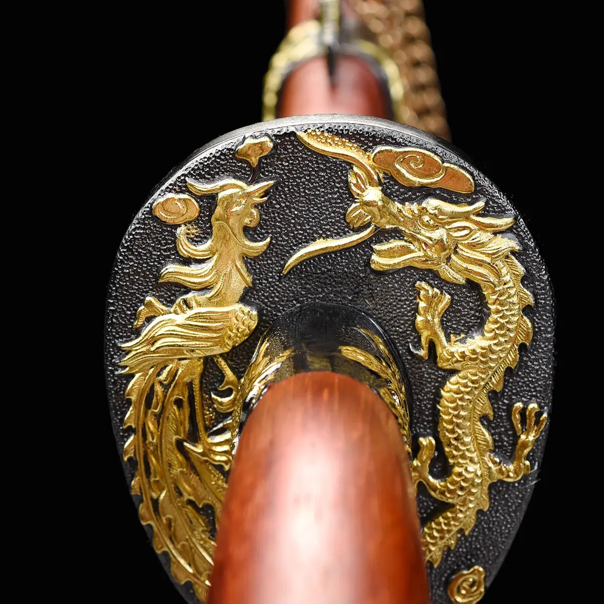 Gros plan frontal sur la garde du sabre chinois Dao LONG FENG, avec motifs dragon et phénix dorés en relief sur fond noir texturé, poignée rouge-brun en bois et lame partiellement visible en arrière-plan.