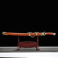 Sabre chinois Dao LONG FENG présenté dans son fourreau rouge-brun en bois, posé horizontalement sur un support en bois devant un fond noir, avec poignée assortie, garde noire, garnitures dorées sculptées et chaîne décorative centrale visibles.