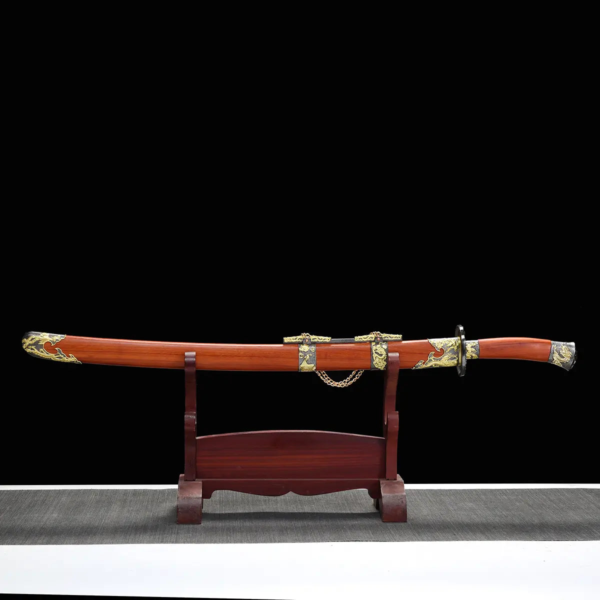 Sabre chinois Dao LONG FENG présenté dans son fourreau rouge-brun en bois, posé horizontalement sur un support en bois devant un fond noir, avec poignée assortie, garde noire, garnitures dorées sculptées et chaîne décorative centrale visibles.