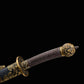 Gros plan sur la poignée brune tressée du sabre chinois Dao LONG WEI, avec pommeau décoré en laiton, garde ronde ornée de dragons et partie du fourreau noir texturé avec garnitures dorées sur fond noir.