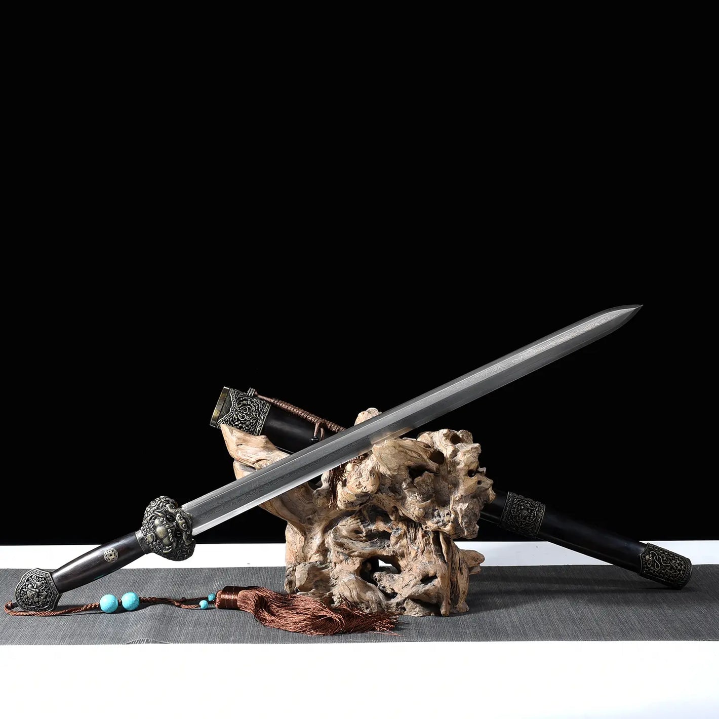 Épée chinoise Jian Longquan présentée sur un support en bois naturel, avec lame droite en acier plié, fourreau sombre type ébène, garde métallique ornementée, pompon brun, perles turquoise, tapis gris et fond noir.