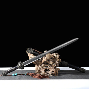 Épée chinoise Jian Longquan présentée sur un support en bois naturel, avec lame droite en acier plié, fourreau sombre type ébène, garde métallique ornementée, pompon brun, perles turquoise, tapis gris et fond noir.