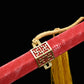 Gros plan sur le fourreau rouge texturé de l’épée chinoise Jian LONGQUAN HONG LONG, avec garniture dorée ajourée, corde tressée jaune, pompon décoratif et fond noir.