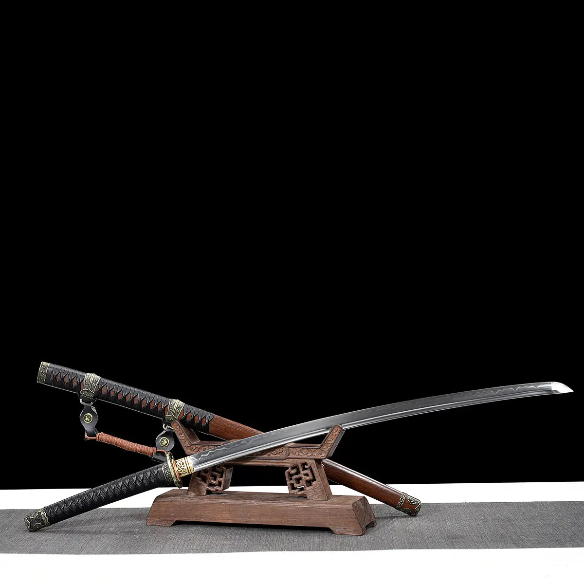 Japanese katana Meiyo displayed crossed on a carved wooden display stand against a solid black background with a light gray surface below. The katana is shown unsheathed, with the polished T10 steel blade crossing above the dark brown wooden saya. The real wavy hamon is visible along the blade edge, while the black tsuka wrapped in black ito over rayskin features antique-style metal fittings. The composition highlights the dynamic crossed presentation, traditional koshirae, and contrast between the reflecti