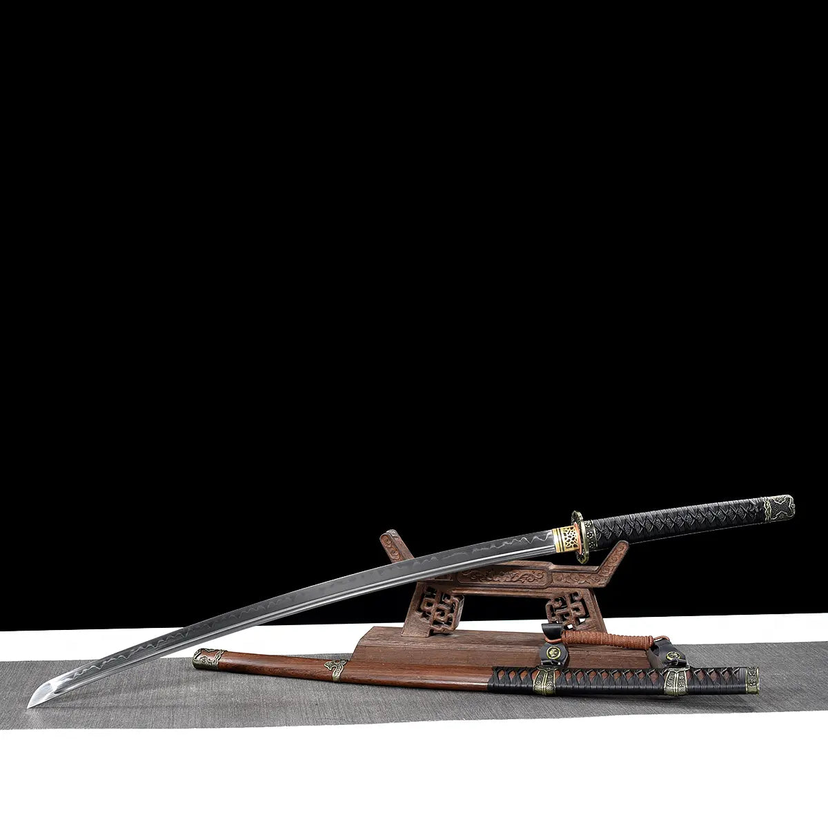 Japanese katana Meiyo displayed diagonally on a carved wooden display stand against a solid black background with a light gray surface below. The polished T10 steel blade is shown unsheathed, extending to the left with a visible wavy hamon along the cutting edge and a defined kissaki tip. The black tsuka wrapped in black ito over rayskin rests on the stand, while the dark brown wooden saya lies below with decorative metal fittings and a sageo cord. The composition emphasizes the blade length, curvature, and