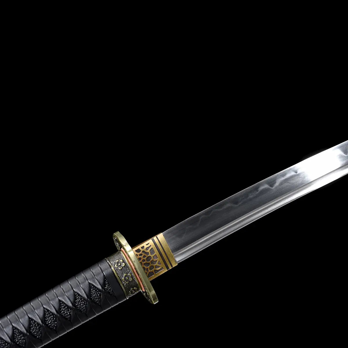Close-up view of the Japanese katana Meiyo on a solid black background, focusing on the base of the T10 steel blade and the transition area between blade and handle. The image highlights the real wavy hamon formed by clay tempering, the decorative brass habaki with cutout pattern, and the round tsuba with antique-style floral details. Part of the black tsuka wrapped in black ito over rayskin is visible, showing the tight wrapping and textured grip, with no scabbard or stand present.
