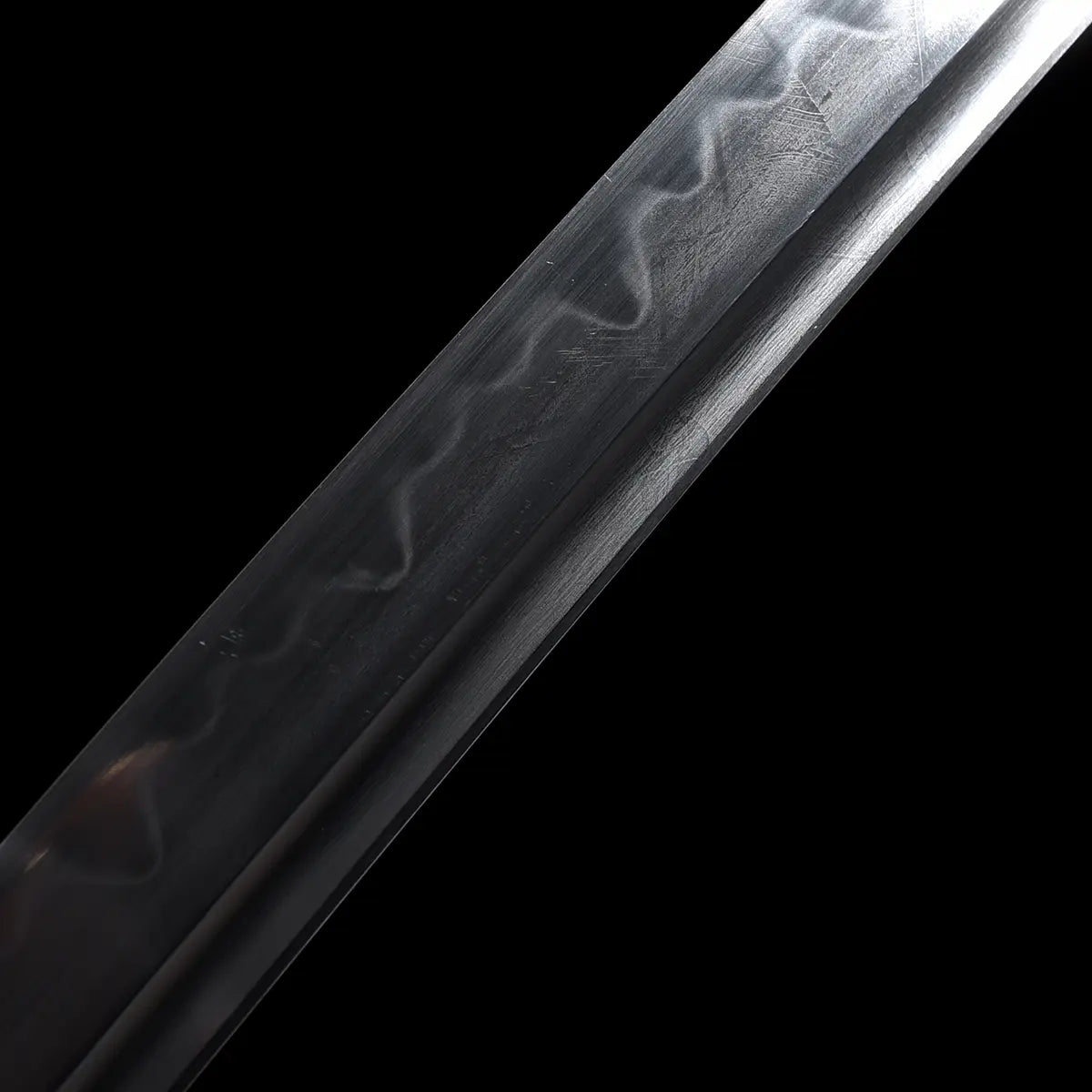 Detailed close-up of the Japanese katana Meiyo blade on a solid black background, focusing on the mid-section of the polished T10 steel blade. The image clearly shows the natural wavy hamon created by traditional clay tempering, running along the cutting edge. Fine surface textures from polishing are visible, along with the clean shinogi line and smooth curvature of the blade. No handle, scabbard, or stand is visible, emphasizing the steel structure, hamon pattern, and contrast between the dark background a