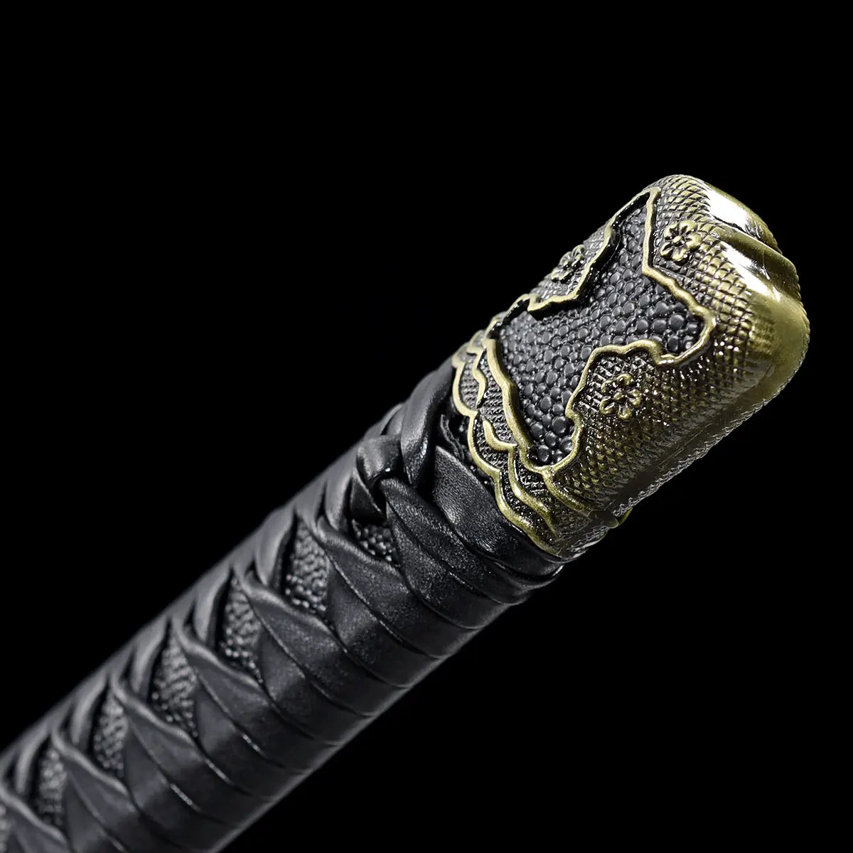 Close-up detail of the Japanese katana Meiyo handle end on a solid black background, focusing on the kashira with antique-style metal finish and floral decorative motifs. The image shows the black ito wrapping tightly braided over black rayskin, with clear diamond pattern texture visible along the tsuka. The metal kashira features engraved details and a slightly aged patina, highlighting the traditional craftsmanship and refined finish of the handle assembly without any blade or scabbard visible.
