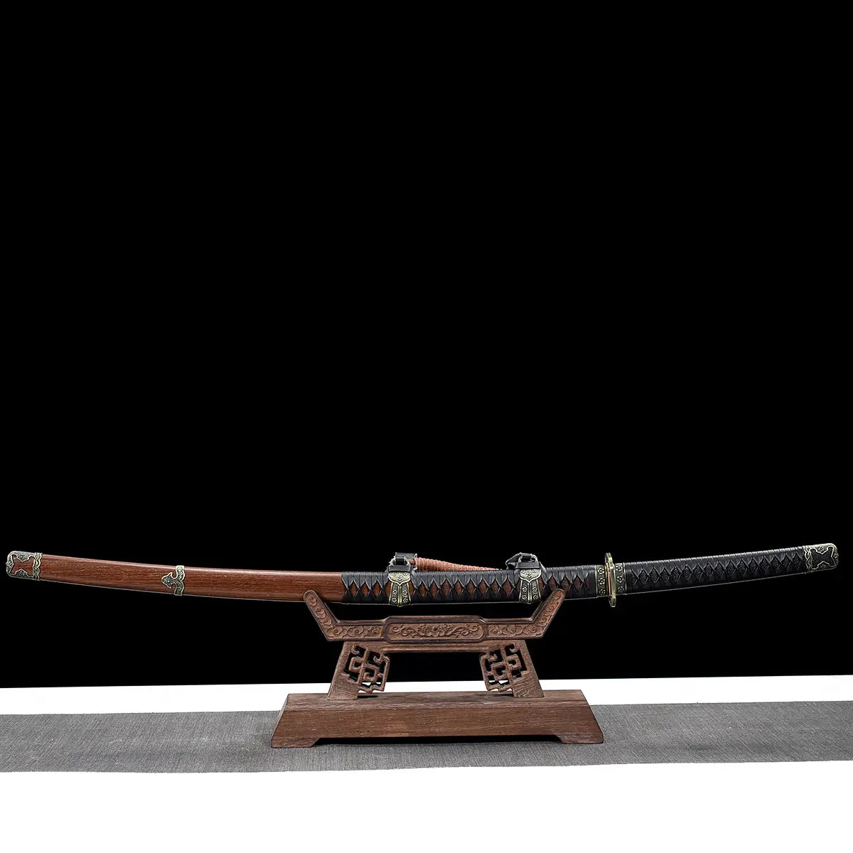 Japanese katana Meiyo displayed horizontally on a carved wooden display stand, set against a solid black background with a light gray surface underneath. The sword is shown fully sheathed, with the dark brown wooden saya resting on the stand and the black tsuka extending to the right. The handle is wrapped in black ito over black rayskin, featuring antique-style metal fittings, while the saya includes decorative metal accents and a sageo cord. The wooden stand has engraved details and supports the katana ev