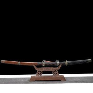 Japanese katana Meiyo displayed horizontally on a carved wooden display stand, set against a solid black background with a light gray surface underneath. The sword is shown fully sheathed, with the dark brown wooden saya resting on the stand and the black tsuka extending to the right. The handle is wrapped in black ito over black rayskin, featuring antique-style metal fittings, while the saya includes decorative metal accents and a sageo cord. The wooden stand has engraved details and supports the katana ev