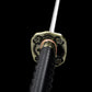 Close-up view of the Japanese katana Meiyo handle and guard on a solid black background, focusing on the detailed tsuba with floral motifs and antique-style metal finish. The image shows the brass habaki fitted tightly at the base of the blade, along with the black tsuka wrapped in black ito over rayskin. The perspective highlights the alignment between the blade, habaki, and tsuba, emphasizing the craftsmanship, traditional koshirae assembly, and contrast between the polished steel blade and the dark handl
