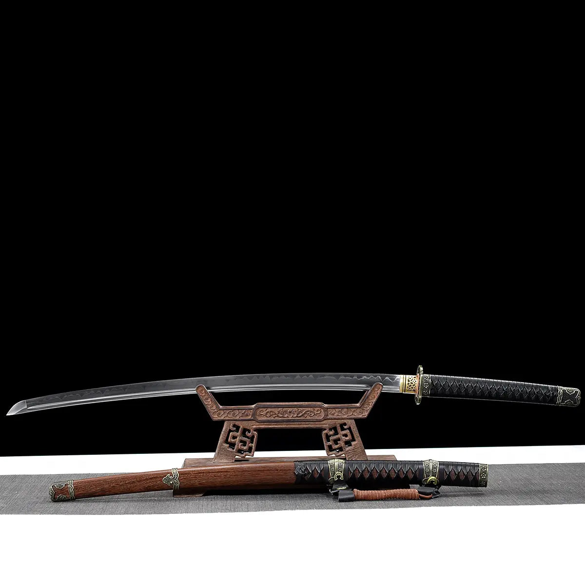 Japanese katana Meiyo displayed unsheathed on a carved wooden display stand, set against a solid black background with a light gray surface below. The polished T10 steel blade is fully visible, showing a real wavy hamon along the cutting edge and a sharp kissaki tip. The black tsuka wrapped in black ito over rayskin extends to the right, while the dark brown wooden saya rests below the stand, partially visible with decorative metal fittings and sageo cord. The composition highlights the full blade profile, 