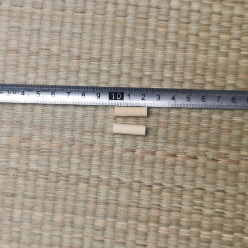 Deux mekugi en bambou clair posés sous une règle métallique graduée en millimètres, repère 10 cm visible, sur un tatami tressé beige ; aucune lame ni fourreau présents. Accessoire mekugi bambou katana, mesure 2–3 cm, HEMA AMHE.

