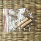 Gros plan d’un petit sachet zip transparent contenant deux mekugi en bambou clair, extrémités rondes, posé sur un tatami tressé beige ; aucun katana, garde ou fourreau visible. Accessoire mekugi bambou katana, HEMA AMHE.
