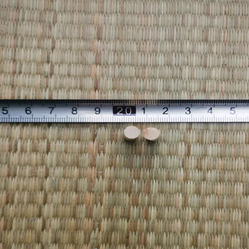 Gros plan de deux mekugi en bambou vus par l’extrémité, section ronde, posés devant une règle métallique graduée avec repère 20 cm, sur tatami tressé beige ; accessoire mekugi bambou katana, HEMA AMHE.
