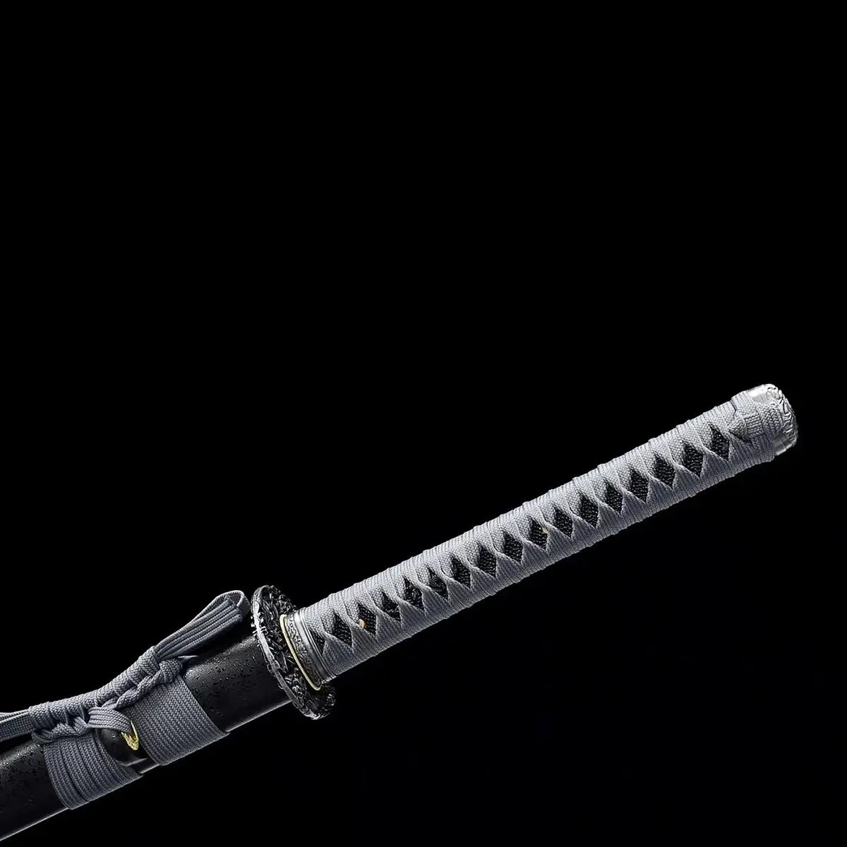 Détail du katana japonais ONI NO KIBA montrant la poignée longue avec tsuka gris tressé laissant apparaître la peau de raie noire, tsuba noire sculptée, fourreau noir laqué avec sageo gris visible, fond noir.

