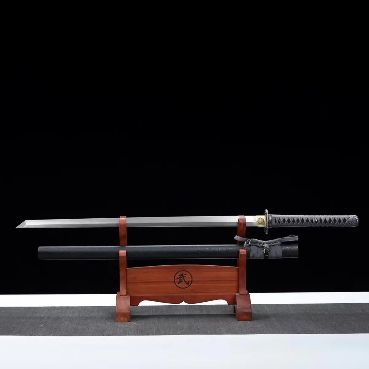 Katana japonais artisanal ONI NO KIBA avec lame dégainée posée horizontalement au-dessus de son fourreau noir laqué sur un support en bois rouge, poignée gris tressé avec peau de raie apparente, tsuba noire sculptée et habaki doré, fond noir avec surface grise.
