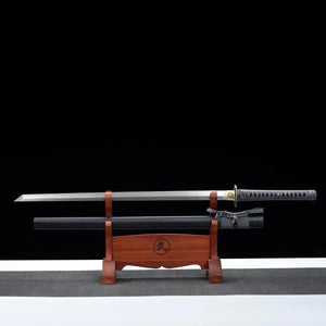 Katana japonais artisanal ONI NO KIBA avec lame dégainée posée horizontalement au-dessus de son fourreau noir laqué sur un support en bois rouge, poignée gris tressé avec peau de raie apparente, tsuba noire sculptée et habaki doré, fond noir avec surface grise.

