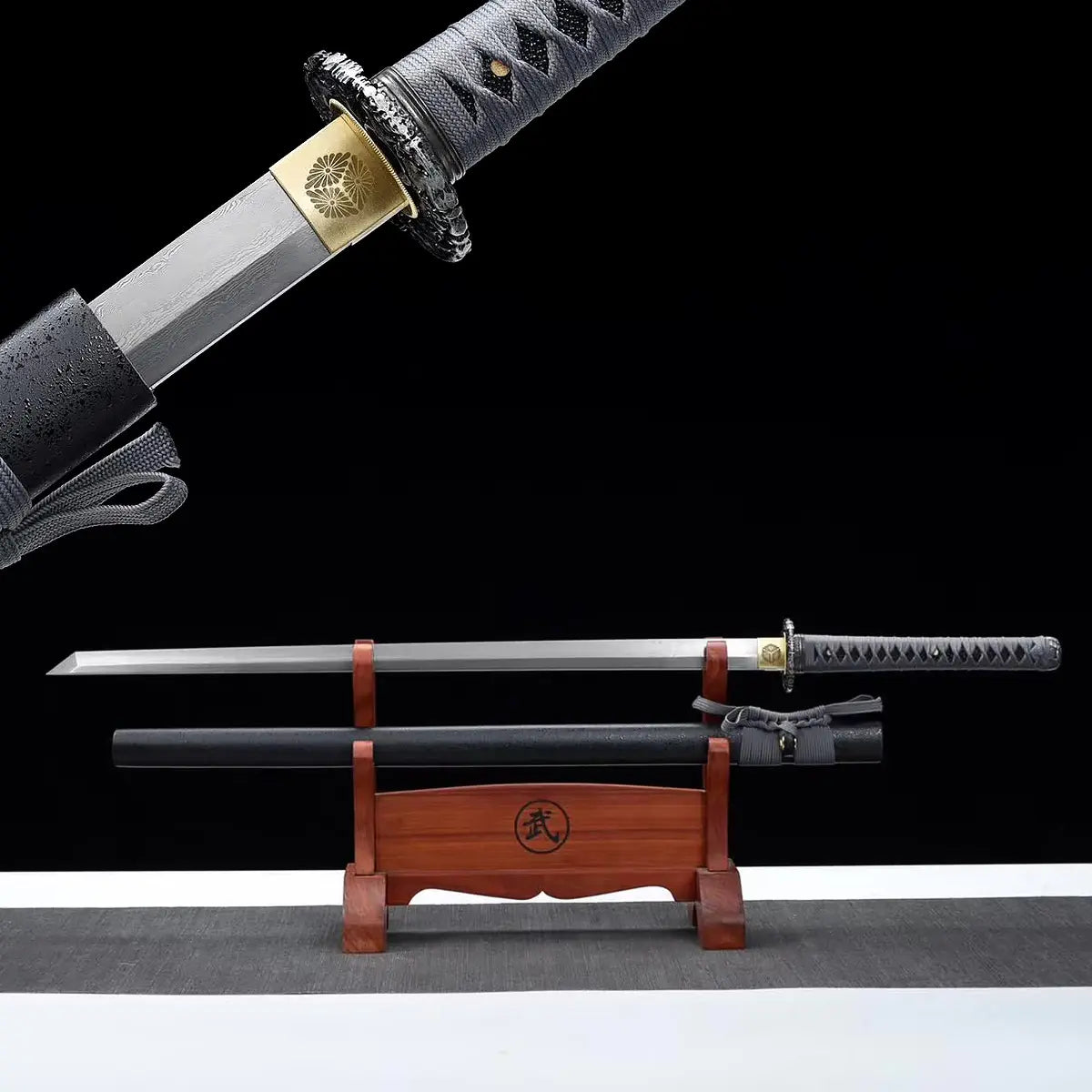Détail du katana japonais ONI NO KIBA montrant la base de la lame en acier avec habaki doré gravé d’un motif floral, tsuba noire sculptée, poignée gris tressé avec peau de raie visible, fourreau noir laqué partiellement visible, fond noir.

