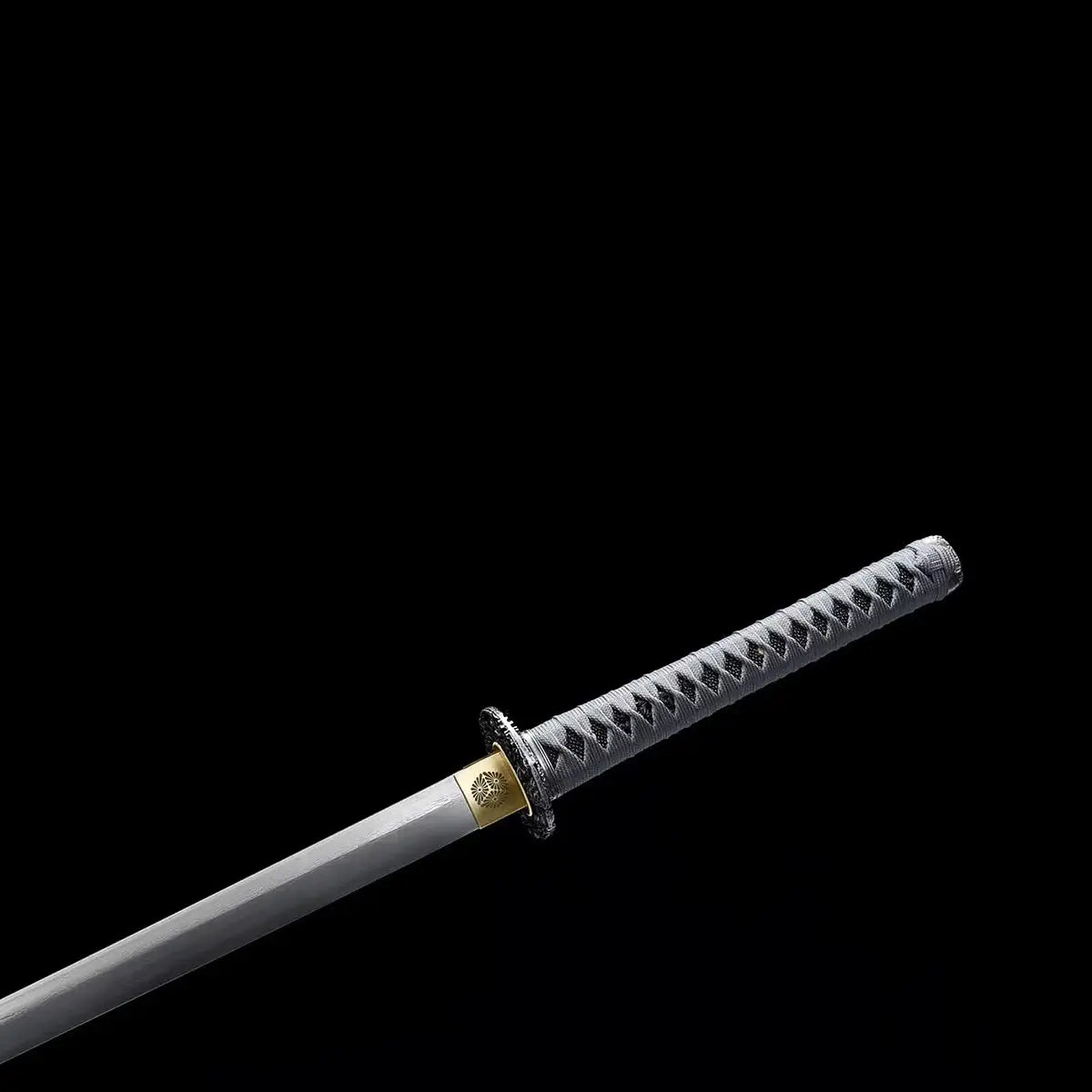 Détail de la poignée du katana japonais ONI NO KIBA avec tsuka gris tressé laissant apparaître la peau de raie, tsuba noire sculptée et habaki doré, lame partiellement visible, sans fourreau, fond noir.
