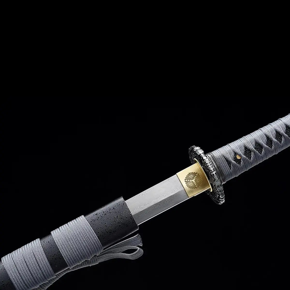 Katana japonais ONI NO KIBA partiellement dégainé montrant la lame sortant d’un fourreau noir laqué avec sageo gris, habaki doré gravé d’un motif floral, tsuba noire sculptée et poignée gris tressé, fond noir.
