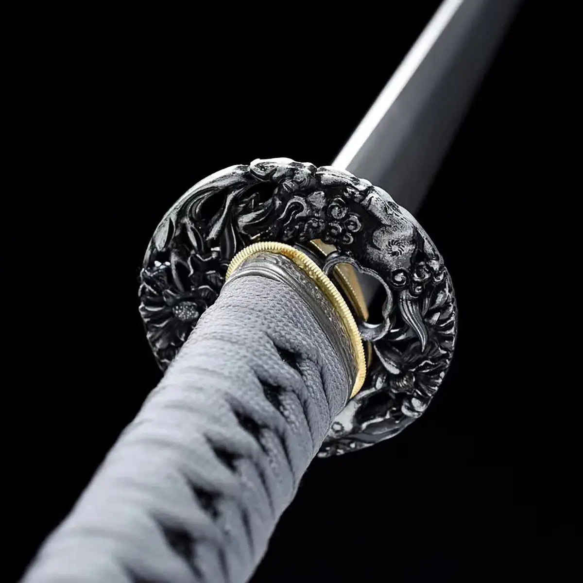 Gros plan du katana japonais ONI NO KIBA montrant une tsuba noire sculptée avec motif de créature, habaki doré, poignée gris tressé avec texture apparente et lame en arrière-plan, fond noir.
