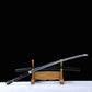Angled horizontal view of a Japanese cosplay katana displayed on a carved wooden stand, photographed against a solid black background with a light-colored floor. The polished steel blade is partially drawn and angled upward, resting above a glossy black lacquered shirasaya scabbard decorated with silver cherry blossom motifs. The sheath is present, no guard is visible, emphasizing a minimalist shirasaya design intended for decorative and cosplay display.
