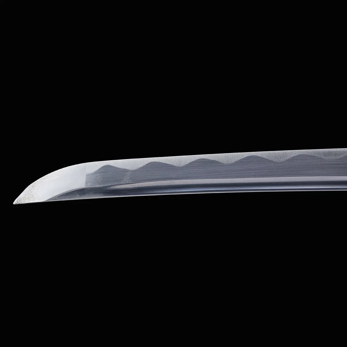 Close-up of the tip of a Japanese cosplay katana blade photographed on a solid black background. The polished steel blade shows a visible wavy hamon line along the edge and a smooth curved kissaki. The scabbard is not visible in this shot, focusing on the blade geometry and surface finish of the shirasaya-style katana.
