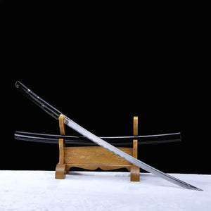 Diagonal view of a Japanese cosplay katana displayed on a carved wooden stand, photographed against a solid black background with a light-colored floor. The polished steel blade is fully visible, resting diagonally above a glossy black lacquered shirasaya scabbard decorated with silver cherry blossom motifs. The sheath is present, no guard is visible, emphasizing a minimalist shirasaya design for decorative and cosplay use.
