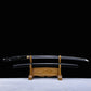 Horizontal view of a Japanese cosplay katana displayed on a carved wooden stand, photographed against a solid black background with a light-colored floor. The katana features a glossy black lacquered shirasaya scabbard decorated with silver cherry blossom motifs, with the polished steel blade fully visible above the saya. The sheath is present, no guard is visible, and the sword is positioned symmetrically for decorative presentation.
