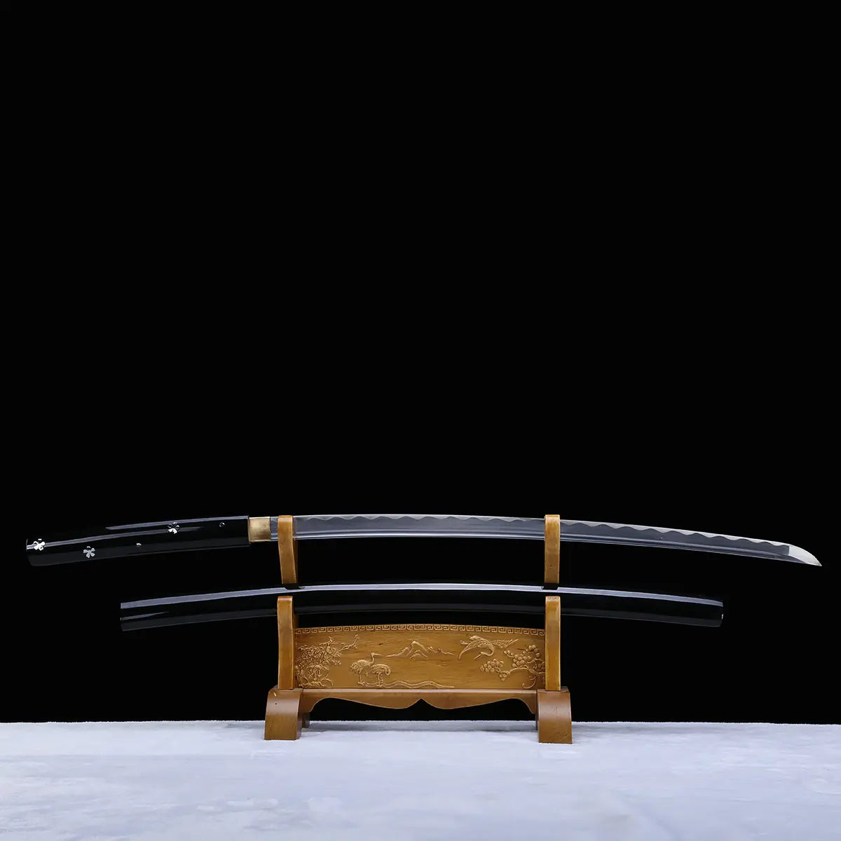 Horizontal view of a Japanese cosplay katana displayed on a carved wooden stand, photographed against a solid black background with a light-colored floor. The katana features a glossy black lacquered shirasaya scabbard decorated with silver cherry blossom motifs, with the polished steel blade fully visible above the saya. The sheath is present, no guard is visible, and the sword is positioned symmetrically for decorative presentation.
